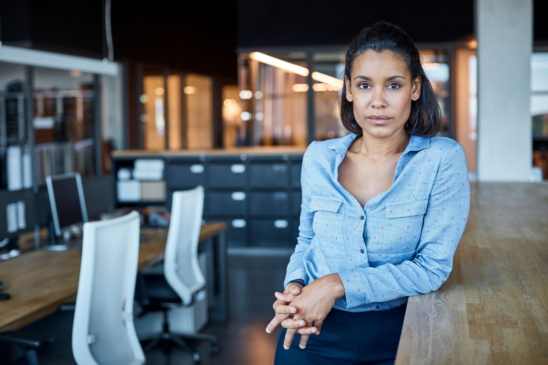 A woman leaning against an office desk, looking thoughtful.